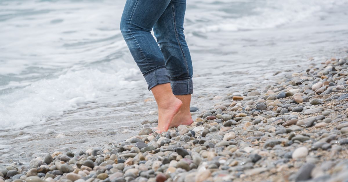 Close-up of a person in jeans walking barefoot along a pebble beach shoreline.