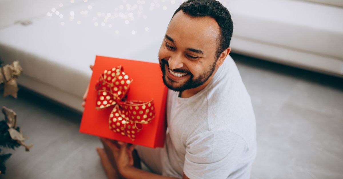 Smiling man holding a red Christmas gift with polka dot ribbon in a cozy living room setting.