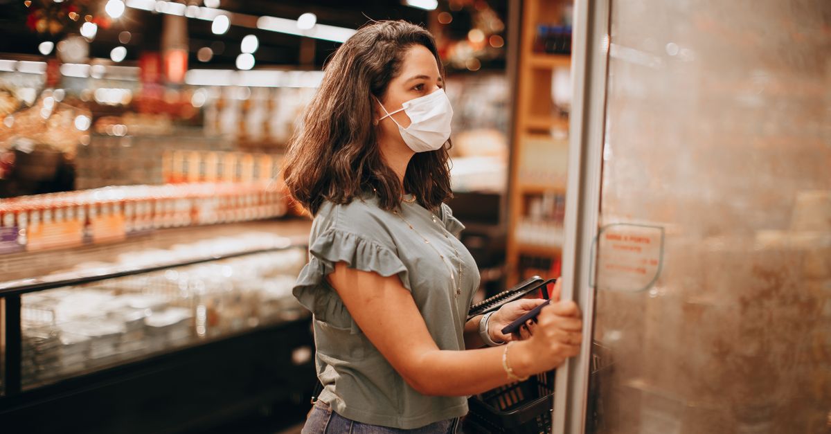 A woman wearing a mask shops in a grocery store, opening a refrigerator.