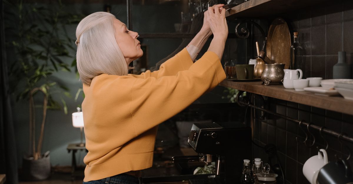 Older woman reaching up to a high kitchen shelf.