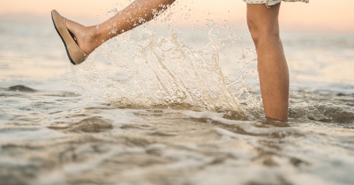 A woman in a floral dress kicks ocean water, creating a splash at sunset.