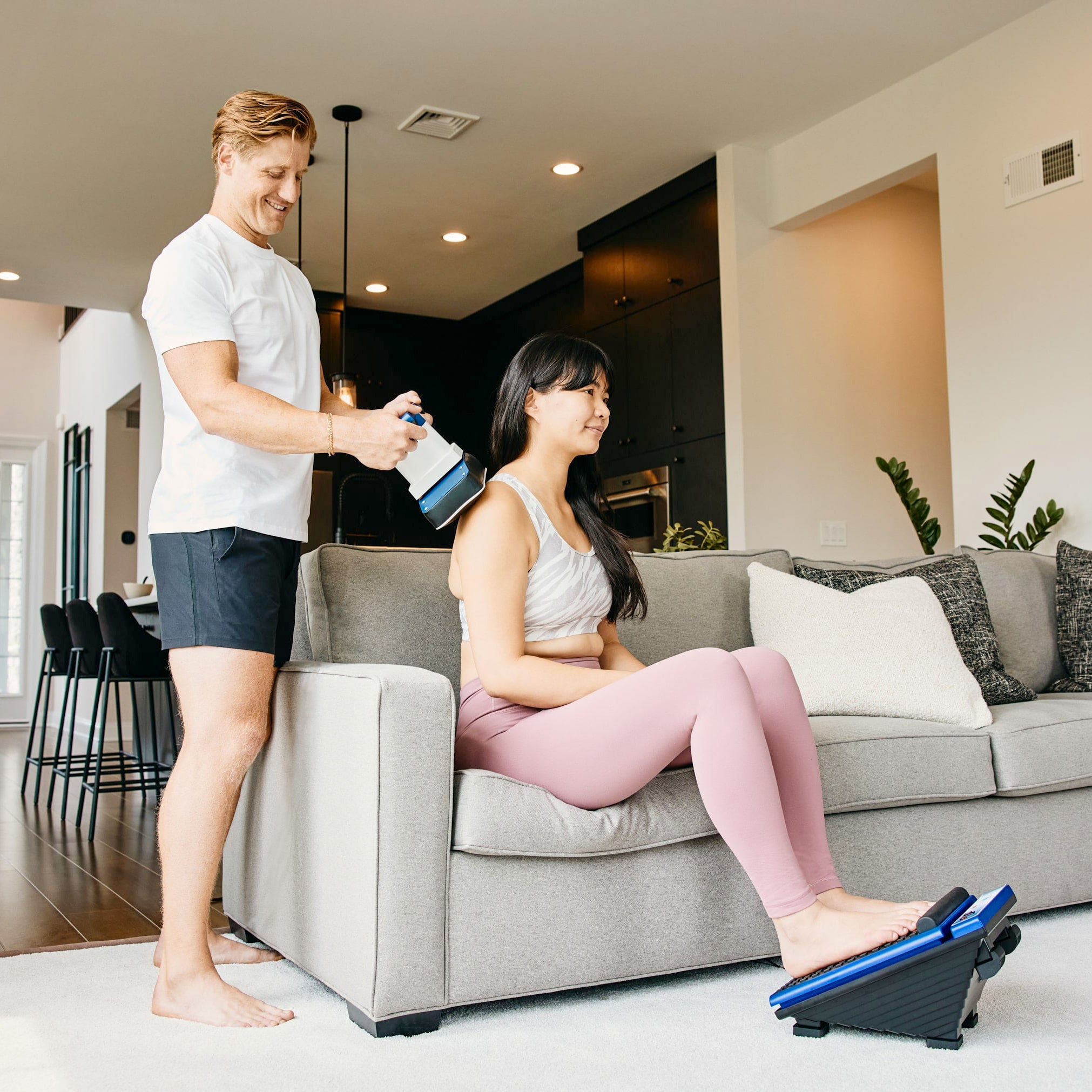 Man using the MedMassager Classic body massager on a woman’s back while she uses the Classic foot massager on a couch at home