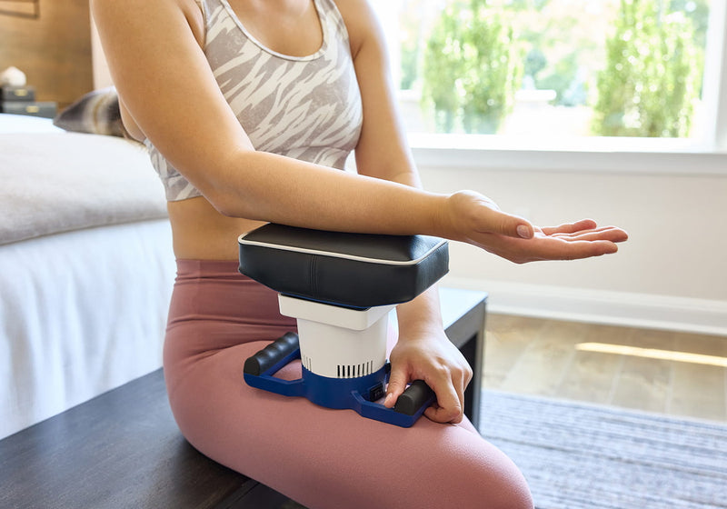 Person sitting indoors rests arm on MedMassager device for blood testing or therapy.