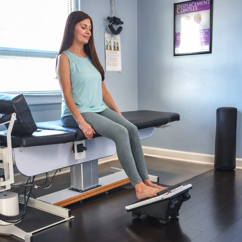 Woman seated with her feet on a MedMassager Foot Massager in a clinic room.