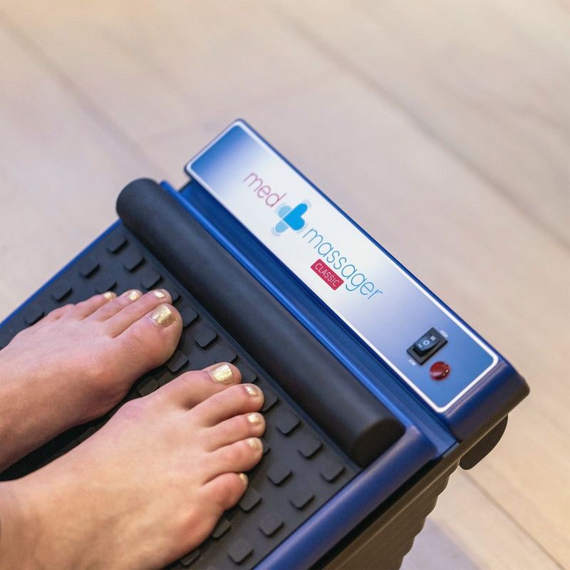 Feet resting on a MedMassager Foot Massager on a wooden floor.