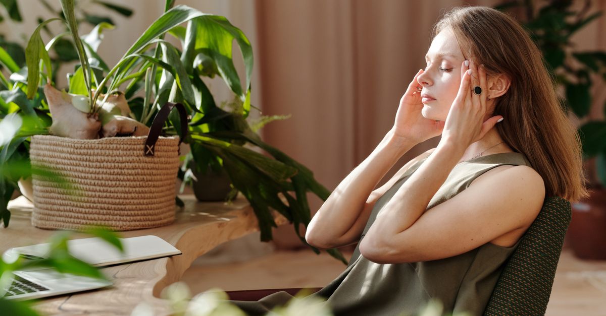 A woman relaxes, massaging her temples among indoor plants in a bright, sunlit room.