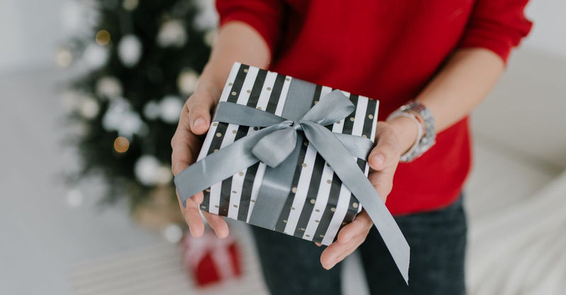 Close-up of a person holding a wrapped gift box with a festive ribbon, in front of a Christmas tree.