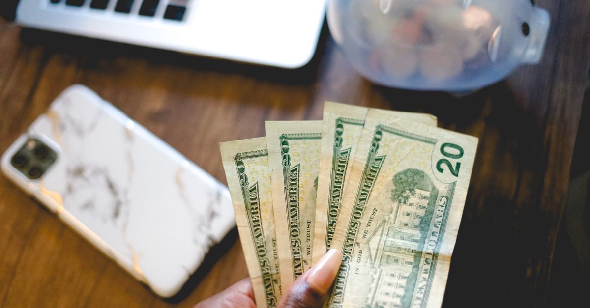 Close-up of a hand holding dollar bills beside a laptop and piggy bank, symbolizing savings and finance.