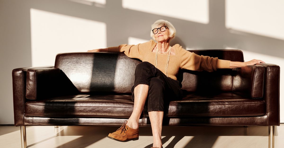Elderly woman with gray hair and eyeglasses relaxing on a leather sofa in a bright room.