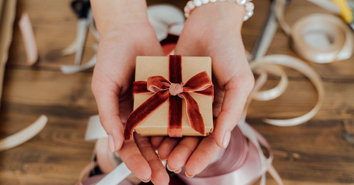 Hands presenting a small gift box wrapped with red velvet ribbon, surrounded by crafting materials.