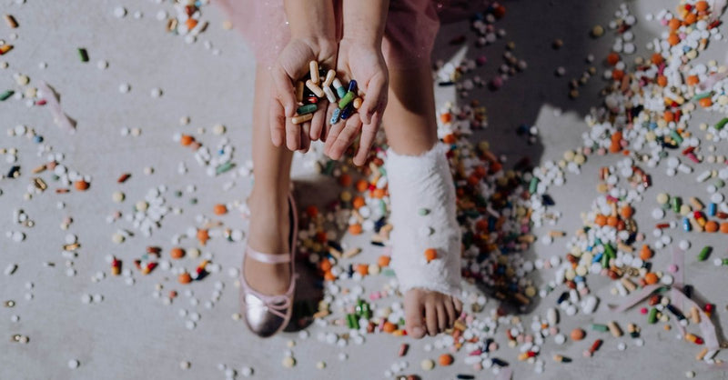 A child with a leg cast sitting among a colorful array of pills indoors, representing medical treatment.
