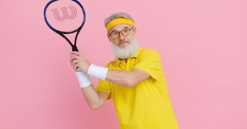 Elderly man in yellow sportswear holding a tennis racket against a vibrant pink background.