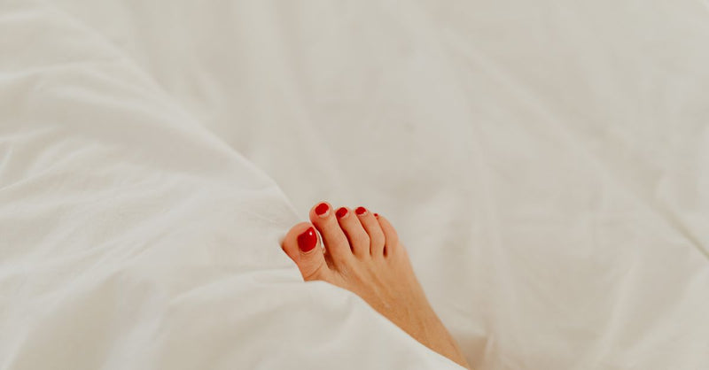 A minimalist and cozy composition of a woman's red-pedicured foot peeking from white bed sheets.