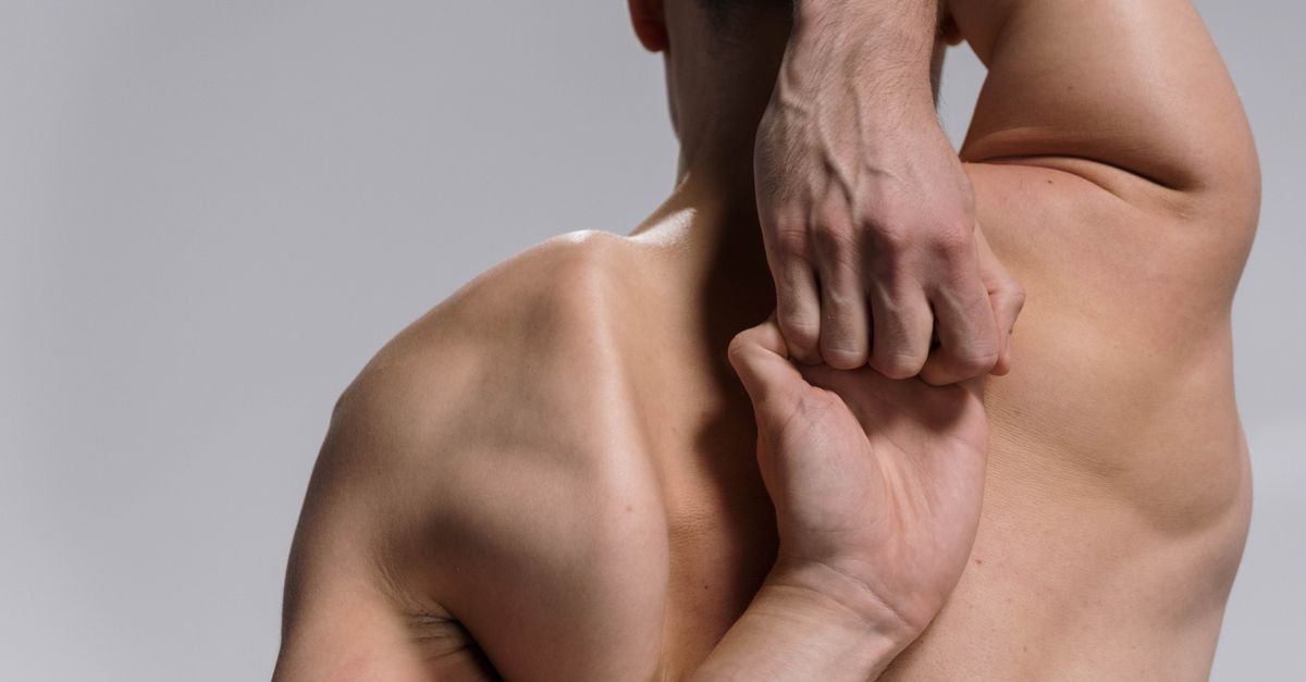 Shirtless muscular man practicing a yoga stretch with hands clasped behind back on a white background.