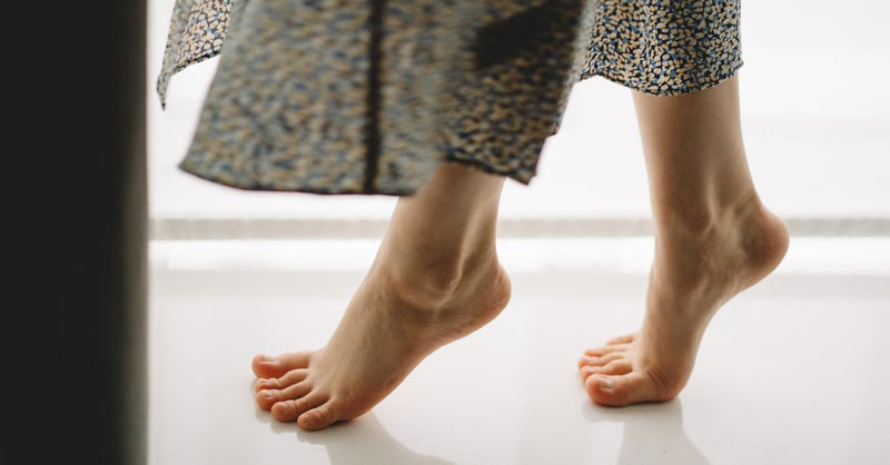 A close-up of a person standing on tiptoes barefoot indoors, wearing a patterned dress.