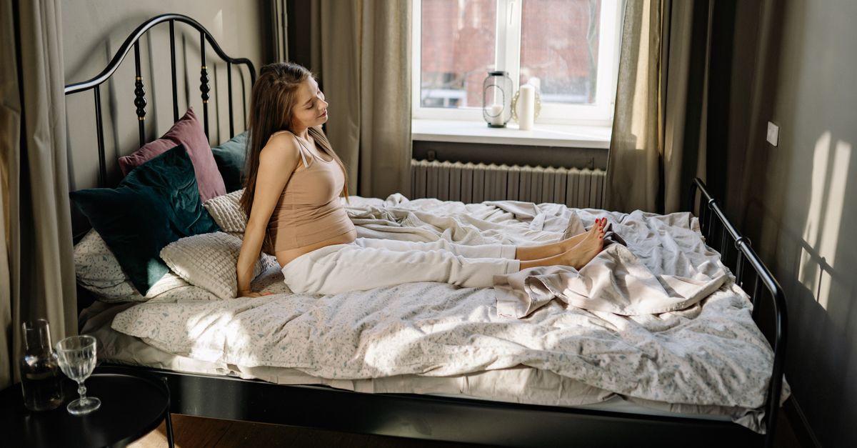 A pregnant woman sitting on a bed in a comfortable, sunlit bedroom.