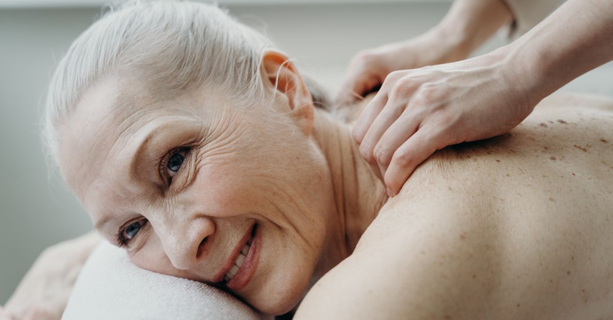 A senior woman with white hair smiles while receiving a therapeutic back massage indoors.