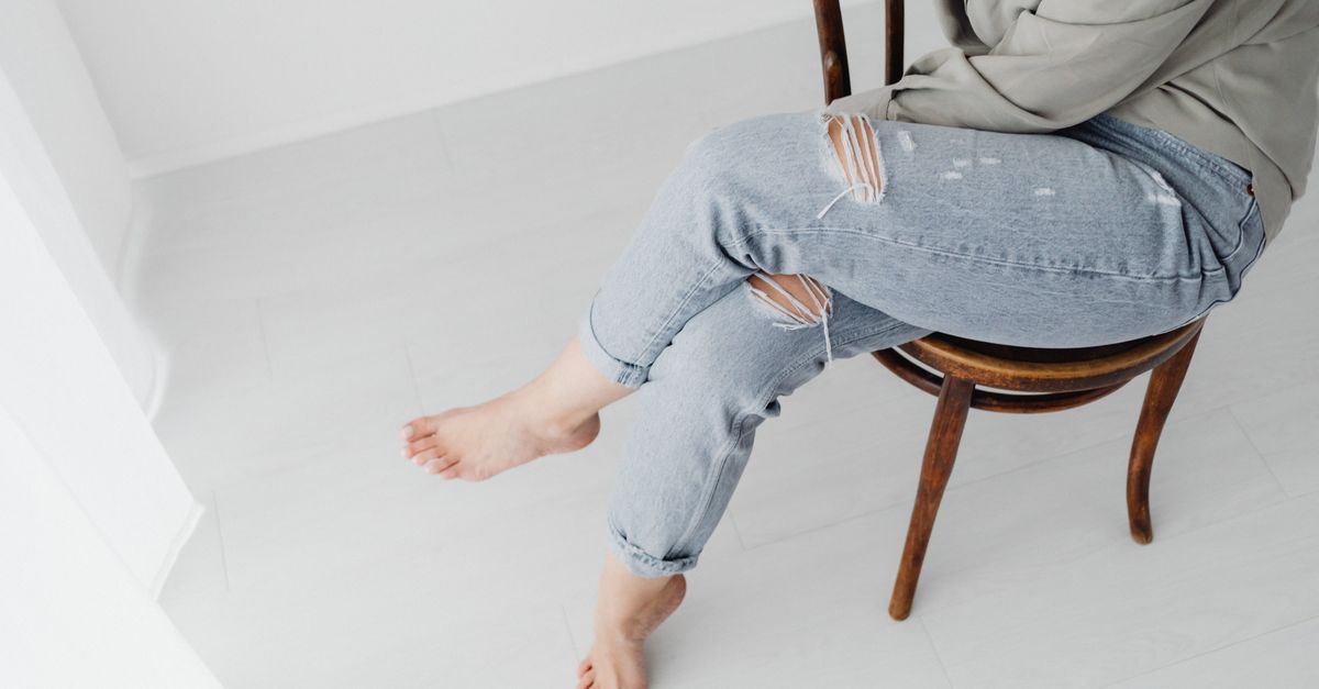 A woman sitting on a wooden chair with crossed legs in a plain, minimal room.