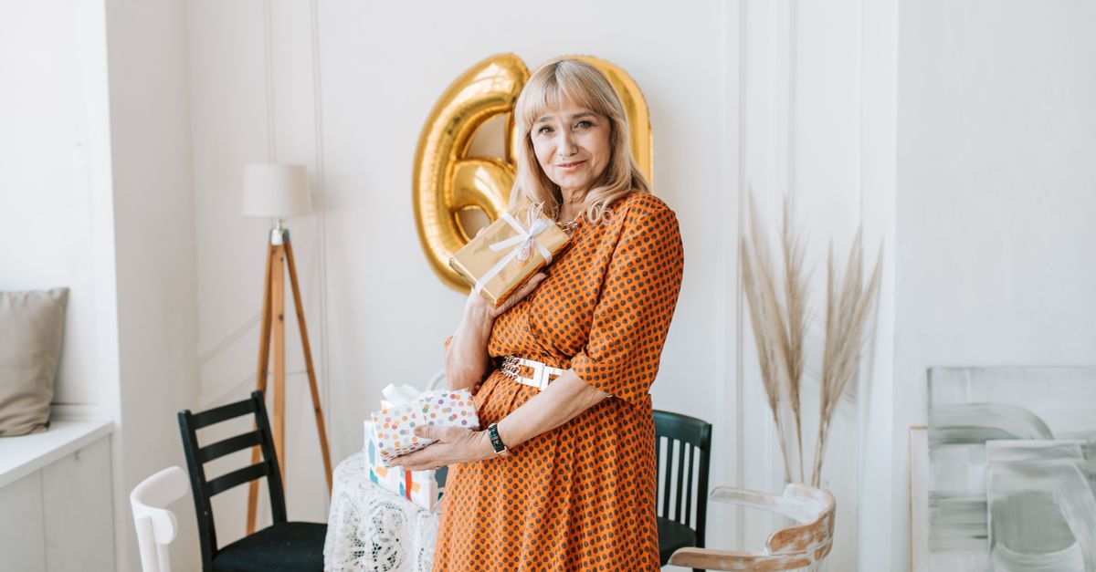 Elderly woman in orange dress holding gifts and smiling, celebrating birthday indoors.