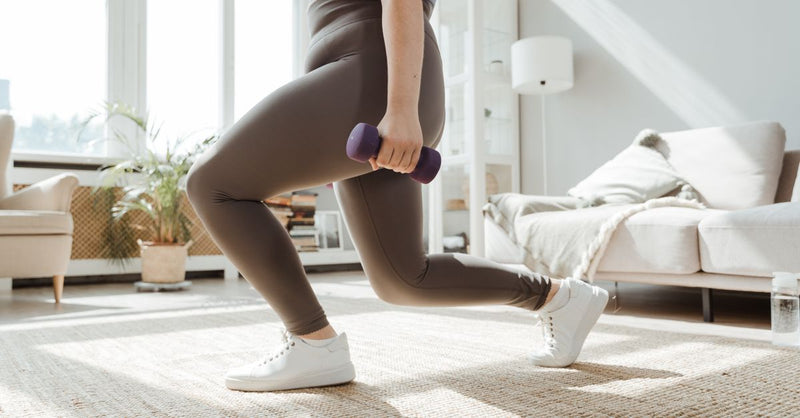 A woman in active wear performs a lunges exercise with dumbbells indoors.