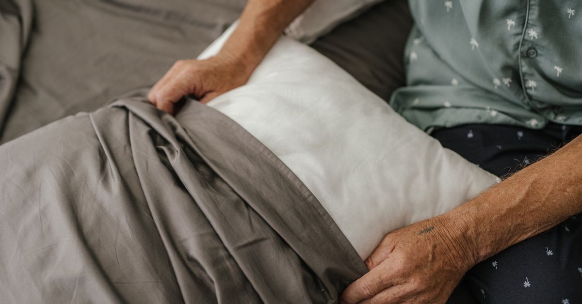 Close-up of a senior adult arranging a bed with grey sheets and a white pillow.