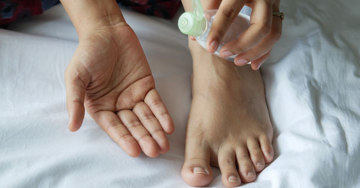 Close-up of hands and foot using a plastic bottle of oil on a bed. Skincare routine concept.