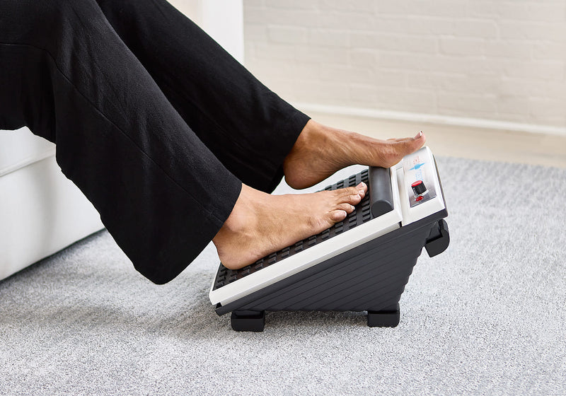 A person uses an electric foot massager while sitting on a sofa, with feet resting on the massager’s textured surface.